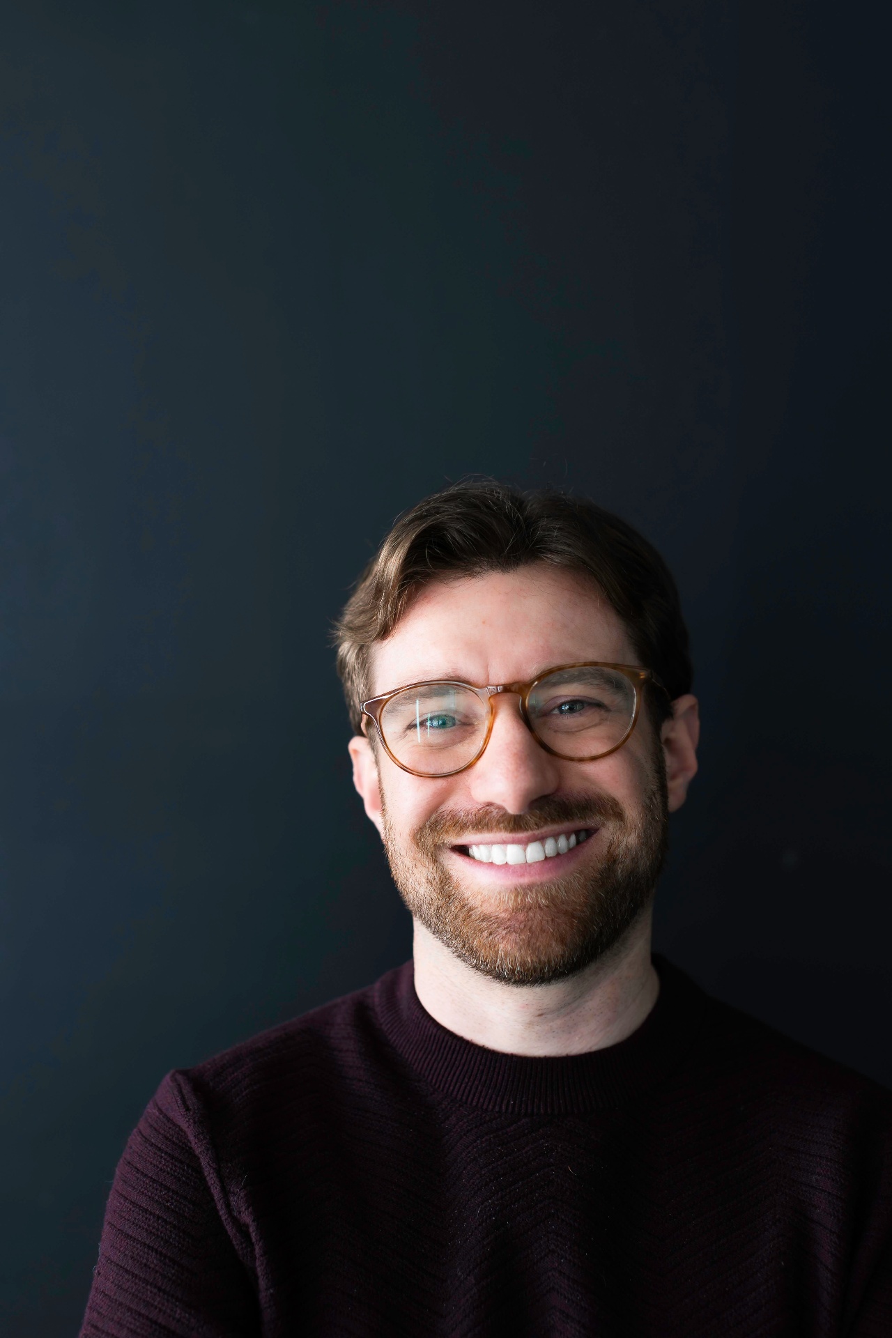Portrait of Charlie Kramer smiling warmly, wearing tortoiseshell glasses and a burgundy sweater against a deep navy background.
